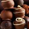 Stack of no-bake chocolate cream cheese truffles on a white plate with a spoon—smooth milk and dark chocolate shells, some cocoa-dusted, others topped with white chocolate swirls and a few purple sugar sprinkles.
