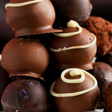 Stack of no-bake chocolate cream cheese truffles on a white plate with a spoon—smooth milk and dark chocolate shells, some cocoa-dusted, others topped with white chocolate swirls and a few purple sugar sprinkles.