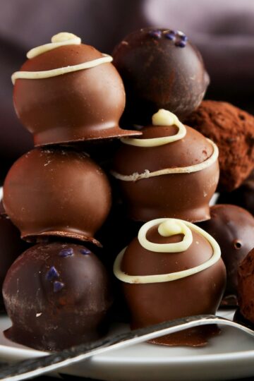 Stack of no-bake chocolate cream cheese truffles on a white plate with a spoon—smooth milk and dark chocolate shells, some cocoa-dusted, others topped with white chocolate swirls and a few purple sugar sprinkles.