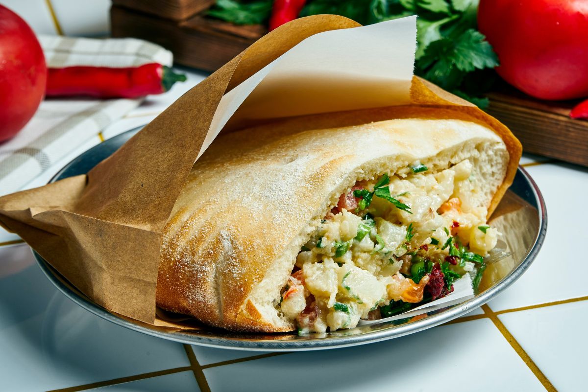 Air fryer cauliflower pita sandwich wrapped in brown parchment, filled with roasted cauliflower, fresh herbs, and creamy tahini sauce inside a lightly toasted pita. Served on a metal plate with tomatoes, chili peppers, and herbs in the background, styled as a healthy cauliflower pita meal.