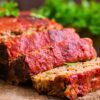 Thick slices of turkey meatloaf rest on a wooden board, showing a tender, veggie-flecked interior and a rich red ketchup glaze on top. Edges are slightly crisp, and fresh parsley sits blurred in the background for a warm, homemade feel.