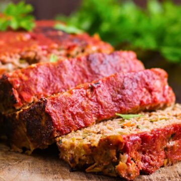 Thick slices of turkey meatloaf rest on a wooden board, showing a tender, veggie-flecked interior and a rich red ketchup glaze on top. Edges are slightly crisp, and fresh parsley sits blurred in the background for a warm, homemade feel.