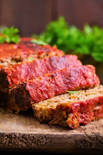 Thick slices of turkey meatloaf rest on a wooden board, showing a tender, veggie-flecked interior and a rich red ketchup glaze on top. Edges are slightly crisp, and fresh parsley sits blurred in the background for a warm, homemade feel.