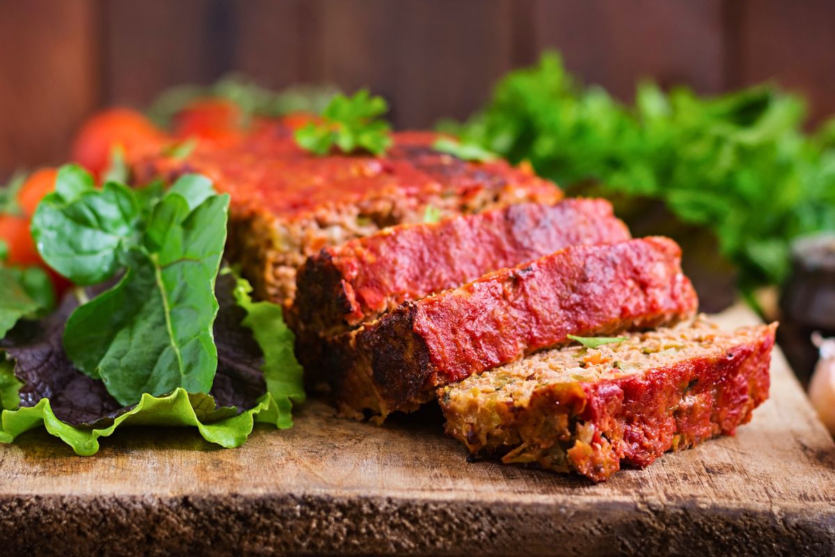 Sliced turkey meatloaf with a glossy red ketchup glaze sits on a rustic wooden board. The tender interior shows herbs and vegetables, and fresh leafy greens and tomatoes rest beside it with a blurred garden-style background.