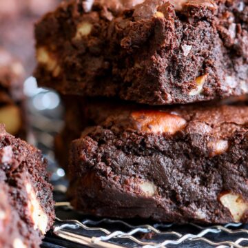 Stacked homemade chocolate walnut brownies on a cooling rack, showing a dense, fudgy texture with visible walnut pieces throughout. Thick brownie squares with rich chocolate crumb and slightly crackled tops, photographed close up on a dark surface.