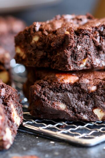 Stacked homemade chocolate walnut brownies on a cooling rack, showing a dense, fudgy texture with visible walnut pieces throughout. Thick brownie squares with rich chocolate crumb and slightly crackled tops, photographed close up on a dark surface.