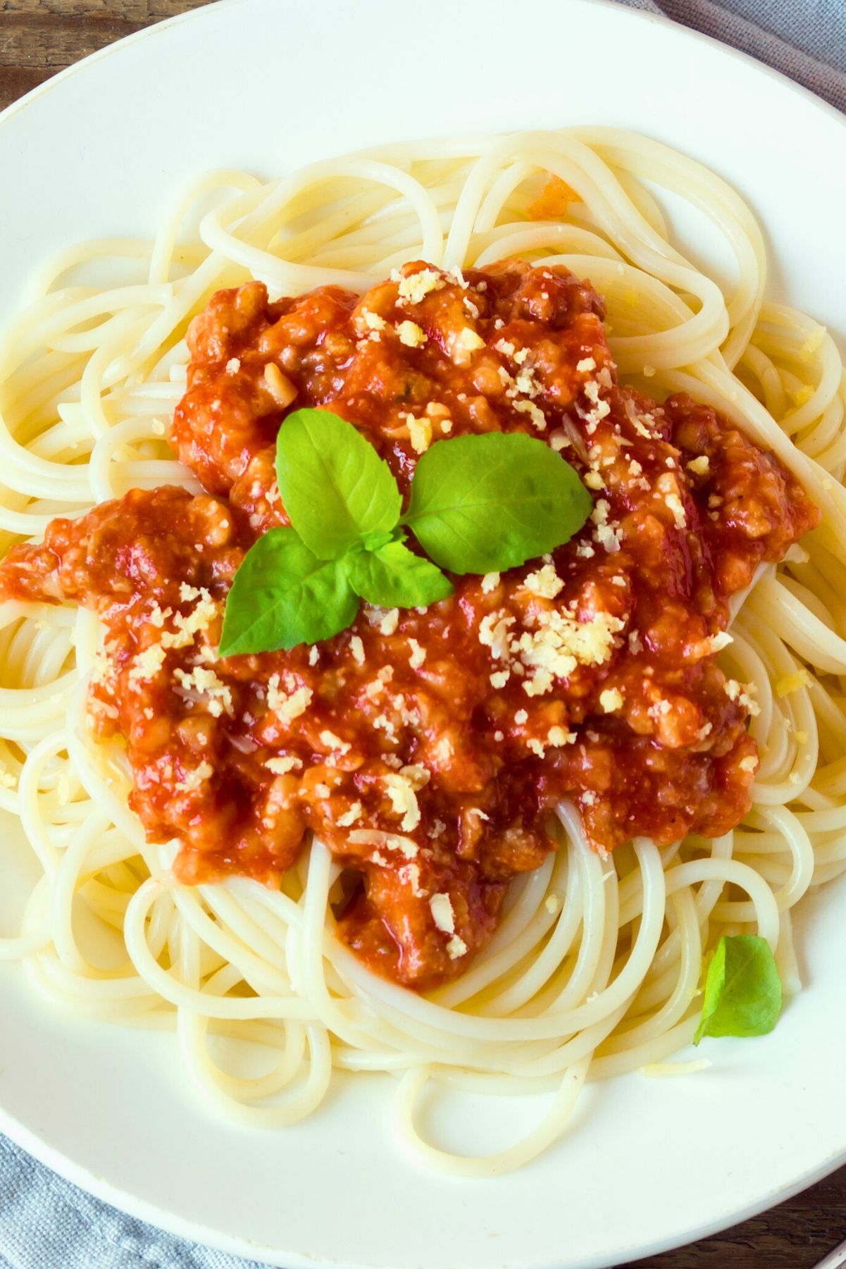 Spaghetti noodles topped with homemade spaghetti sauce made with ground turkey, chunky tomato meat sauce, grated Parmesan, and fresh basil leaves. Served on a white plate on a rustic wooden table with a neutral cloth napkin.