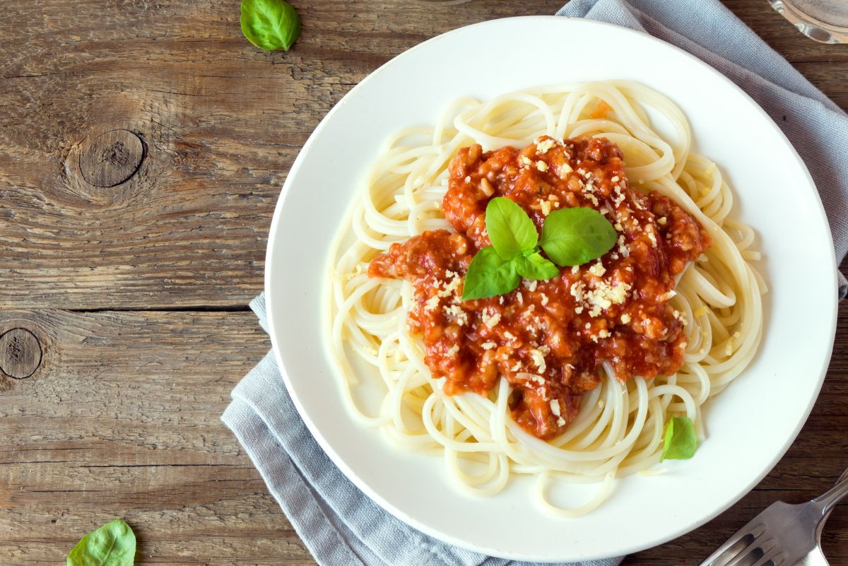 Overhead view of spaghetti topped with homemade spaghetti sauce made with ground turkey, chunky tomato meat sauce, grated Parmesan, and fresh basil. Served on a white plate on a rustic wooden table with a gray cloth napkin and scattered basil leaves.