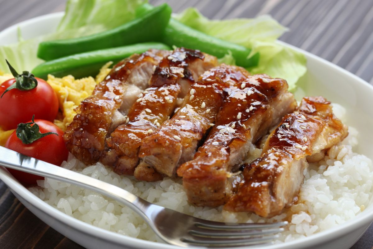 Glazed teriyaki chicken sliced over steamed white rice, sprinkled with sesame seeds. Fresh sugar snap peas, lettuce, scrambled egg, and two bright cherry tomatoes sit alongside, with a silver fork resting at the edge of the bowl.