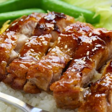 Glazed teriyaki chicken sliced over steamed white rice, topped with toasted sesame seeds. Crisp sugar snap peas sit in the back, adding bright green color. A silver fork rests at the edge of the bowl.