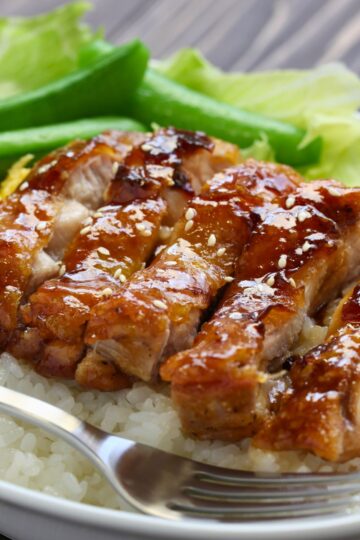 Glazed teriyaki chicken sliced over steamed white rice, topped with toasted sesame seeds. Crisp sugar snap peas sit in the back, adding bright green color. A silver fork rests at the edge of the bowl.