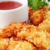 Golden baked coconut shrimp with a crispy toasted coconut coating, served on a white plate beside a small white ramekin of red Thai chili dipping sauce. Fresh parsley garnish adds a pop of green. Close-up food photography highlighting crunchy texture and golden color.