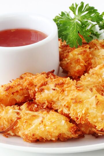 Golden baked coconut shrimp with a crispy toasted coconut coating, served on a white plate beside a small white ramekin of red Thai chili dipping sauce. Fresh parsley garnish adds a pop of green. Close-up food photography highlighting crunchy texture and golden color.