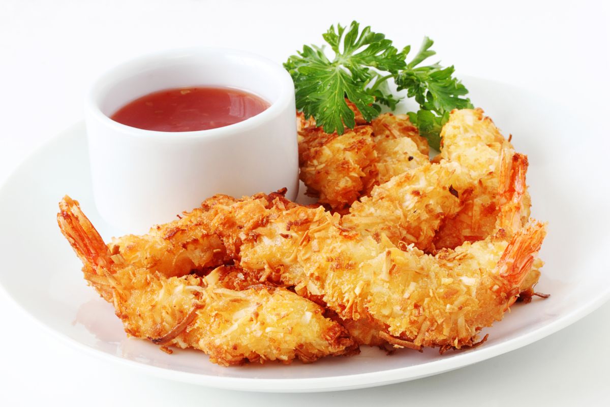 Golden baked coconut shrimp with a crispy toasted coconut coating, arranged on a white plate with a small white ramekin of red Thai chili dipping sauce. Garnished with fresh parsley, highlighting crunchy texture and golden color in clean, bright food photography.