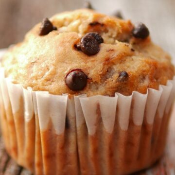 Close-up of a chocolate chip banana mini muffin in a white paper liner on weathered wood, golden top studded with melty chocolate chips.