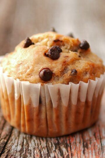 Close-up of a chocolate chip banana mini muffin in a white paper liner on weathered wood, golden top studded with melty chocolate chips.