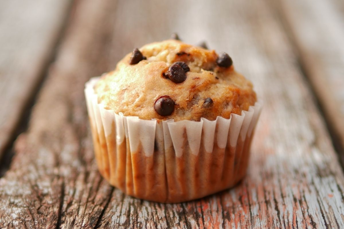 Close-up of a chocolate chip banana mini muffin in a white paper liner on a weathered wooden surface, golden-brown top studded with chocolate chips.
