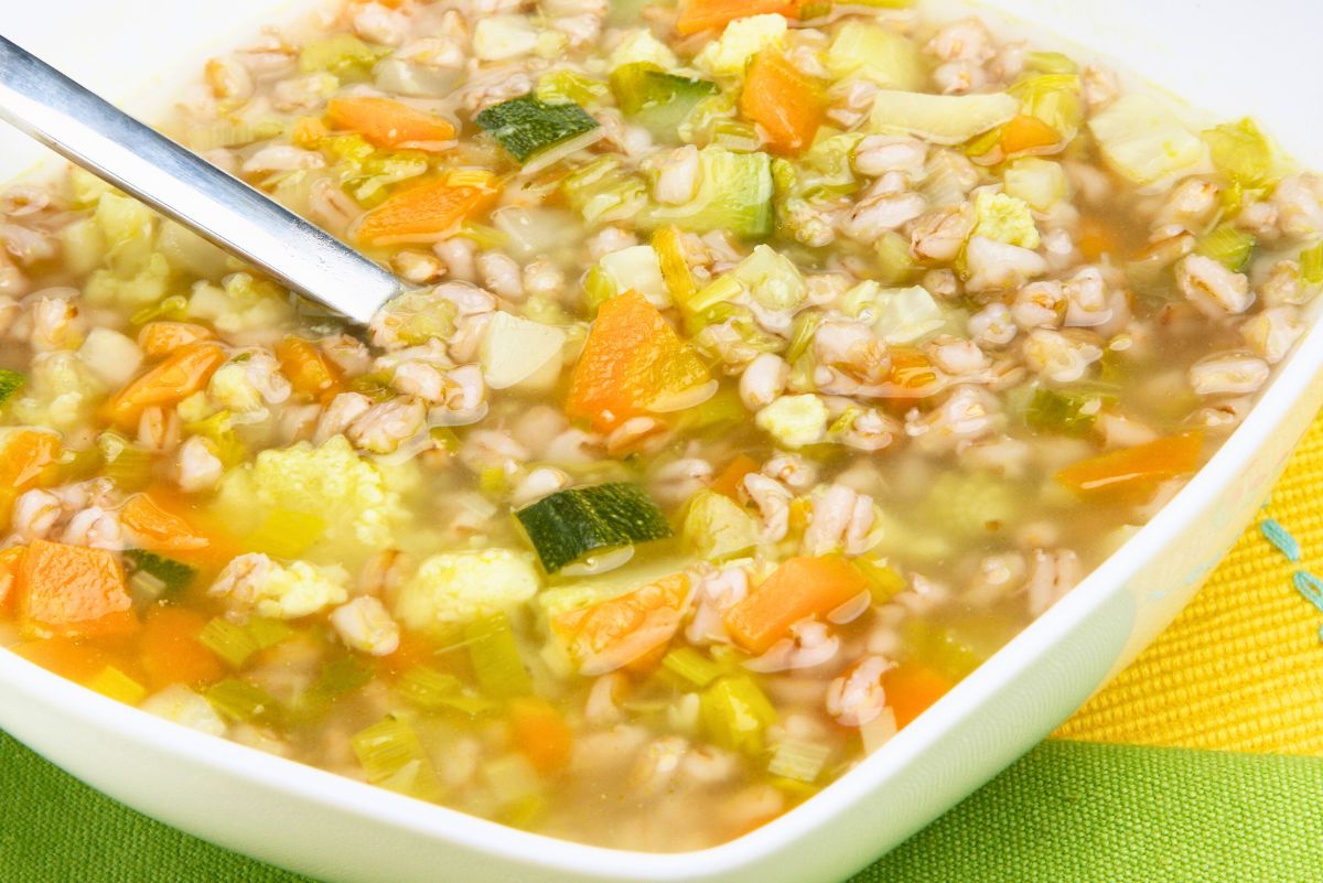 Bowl of vegetable and barley soup with pearl barley, carrots, zucchini, celery, cabbage, and cauliflower in a light golden broth. A metal spoon rests in the soup, with the white bowl sitting on a green kitchen towel, showing a hearty, chunky texture.