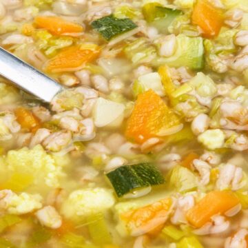 Close-up of vegetable and barley soup in a white bowl with pearl barley, carrots, zucchini, celery, cabbage, and cauliflower in a light golden broth. A metal spoon rests in the soup, showing a hearty, chunky texture and colorful vegetables.