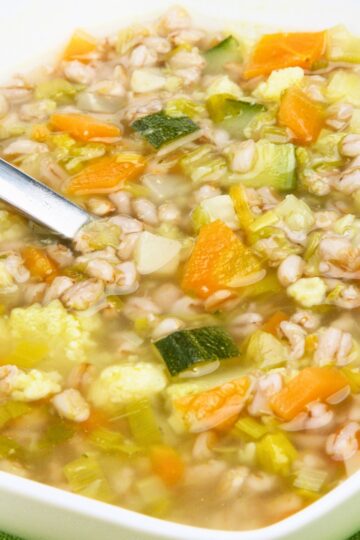 Close-up of vegetable and barley soup in a white bowl with pearl barley, carrots, zucchini, celery, cabbage, and cauliflower in a light golden broth. A metal spoon rests in the soup, showing a hearty, chunky texture and colorful vegetables.