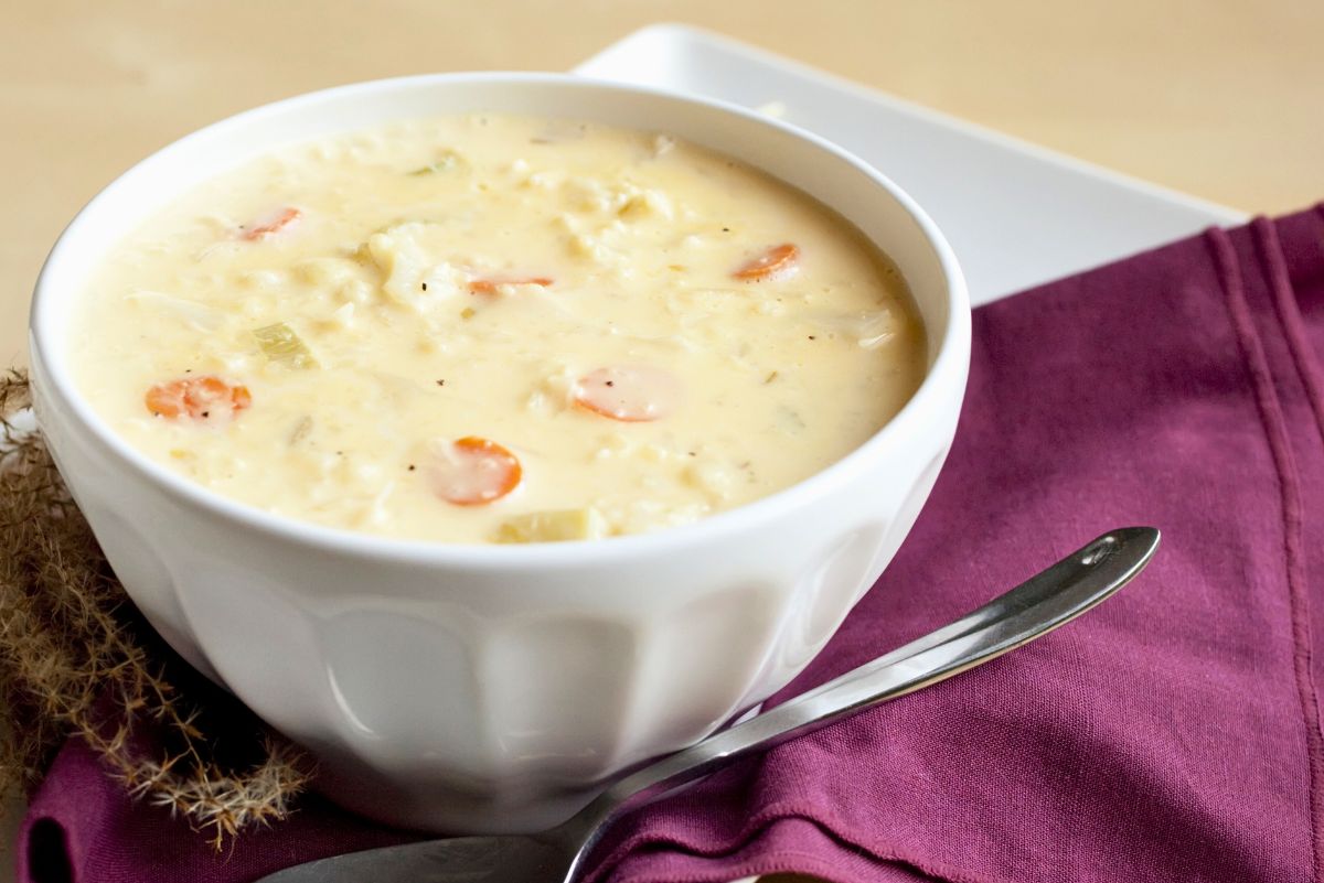Creamy cauliflower cheese soup with carrots served in a white bowl on a white tray. Thick, pale yellow soup with visible cauliflower pieces and carrot slices. Bowl sits on a purple napkin with a silver spoon beside it, styled for cozy comfort food photography.