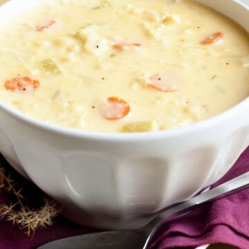 Close-up of creamy cauliflower cheese soup with sliced carrots served in a white bowl. Thick, pale yellow soup with visible cauliflower pieces and carrot rounds, set on a purple napkin with a spoon on a white tray. Cozy, comforting soup presentation with soft lighting.