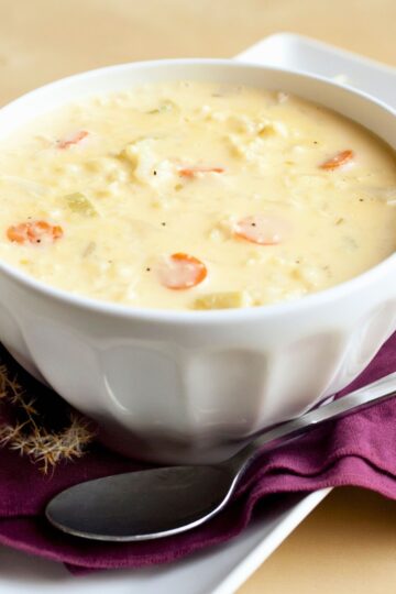 Close-up of creamy cauliflower cheese soup with sliced carrots served in a white bowl. Thick, pale yellow soup with visible cauliflower pieces and carrot rounds, set on a purple napkin with a spoon on a white tray. Cozy, comforting soup presentation with soft lighting.
