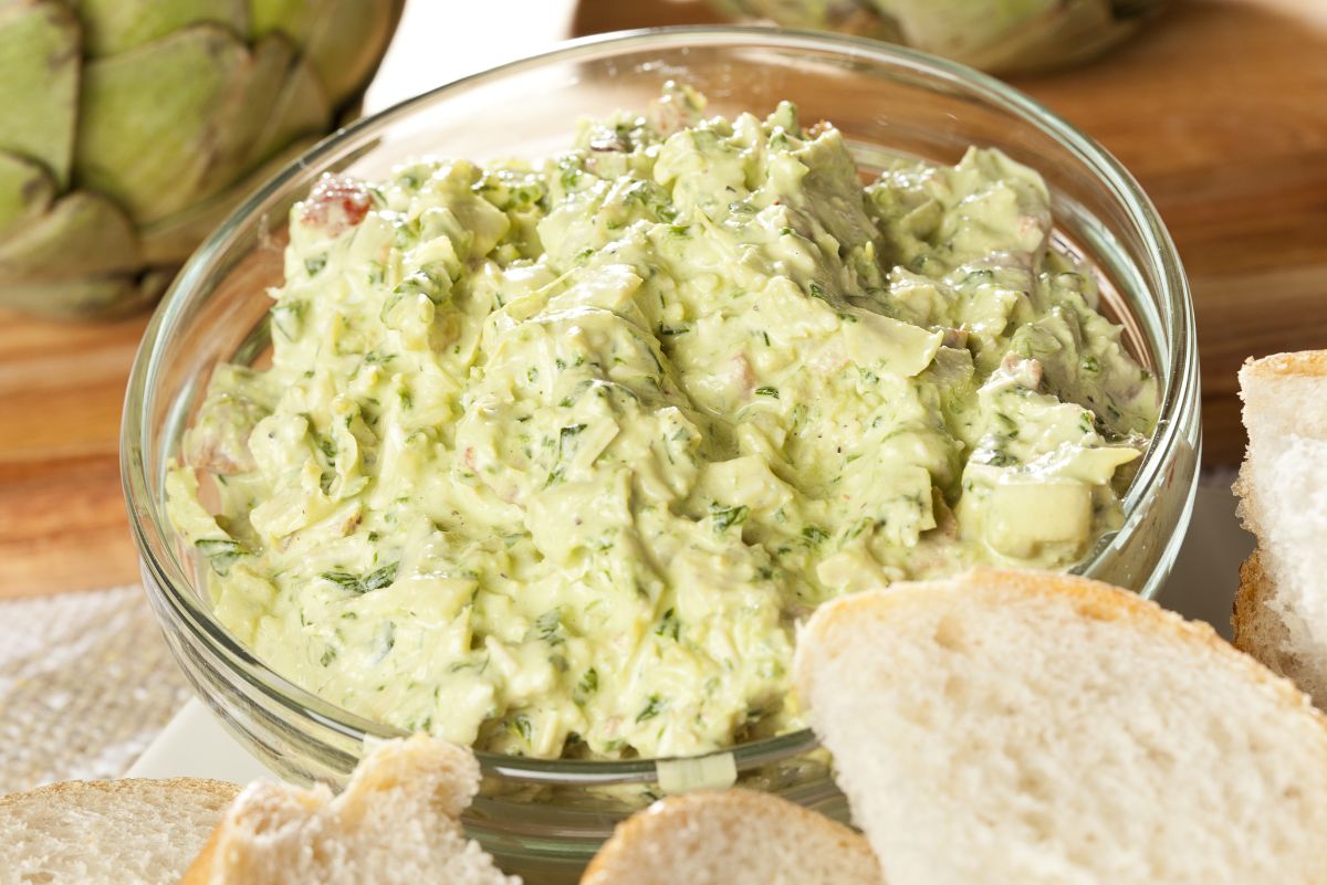 Creamy Greek yogurt artichoke dip in a clear glass bowl, pale green and thick with chopped artichokes and fresh herbs. Served with sliced bread for dipping, with whole artichokes in the background on a wooden surface.