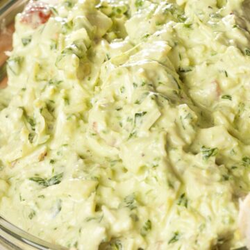 Creamy Greek yogurt artichoke dip in a clear glass bowl, thick and pale green with chopped artichokes, herbs, and small vegetable pieces. Close-up view highlights the smooth, slightly chunky texture, with pieces of bread visible along the edge for dipping.