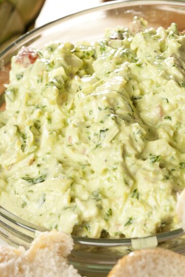 Creamy Greek yogurt artichoke dip in a clear glass bowl, thick and pale green with chopped artichokes, herbs, and small vegetable pieces. Close-up view highlights the smooth, slightly chunky texture, with pieces of bread visible along the edge for dipping.