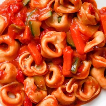 Cheese tortellini tossed in rich tomato sauce with sliced zucchini and tender tomato pieces, served in a white bowl. Close-up view highlights the soft stuffed pasta, glossy red sauce, and green zucchini for a hearty crockpot pasta dinner.