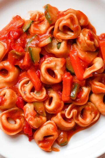 Cheese tortellini tossed in rich tomato sauce with sliced zucchini and tender tomato pieces, served in a white bowl. Close-up view highlights the soft stuffed pasta, glossy red sauce, and green zucchini for a hearty crockpot pasta dinner.