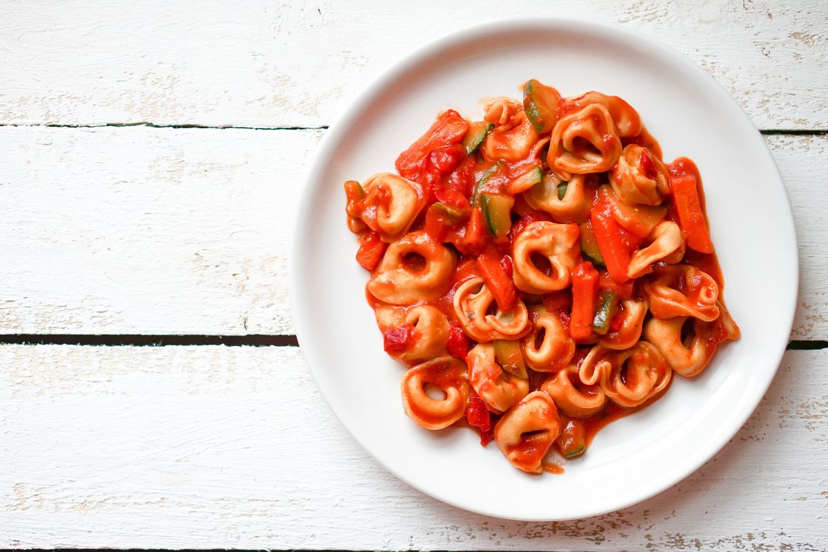 Top-down view of cheese tortellini in red tomato sauce with sliced zucchini and tender vegetables, served on a white plate over a rustic white wood surface. Bright, hearty crockpot pasta dish with visible stuffed tortellini and glossy sauce.