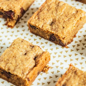 Close-up of easy chocolate chip dessert bars cut into squares, showing a golden baked top and soft interior with melted chocolate chips. Bars are arranged on a light plate with a small gold triangle pattern, highlighting a simple WW friendly dessert bar texture.