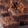 Stack of chocolate mini brownie cupcakes in dark brown paper liners, one topped with a cut brownie piece showing a moist, fudgy center. Light dusting of powdered sugar on a gray surface with soft lighting highlighting the rich chocolate texture.