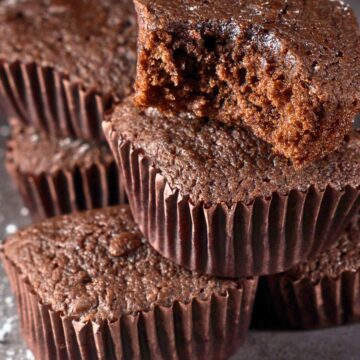 Stack of chocolate mini brownie cupcakes in dark brown paper liners, one topped with a cut brownie piece showing a moist, fudgy center. Light dusting of powdered sugar on a gray surface with soft lighting highlighting the rich chocolate texture.