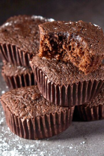 Stack of chocolate mini brownie cupcakes in dark brown paper liners, one topped with a cut brownie piece showing a moist, fudgy center. Light dusting of powdered sugar on a gray surface with soft lighting highlighting the rich chocolate texture.