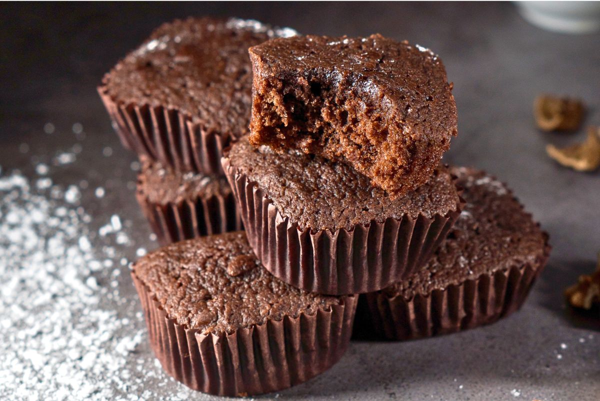 Chocolate mini brownie cupcakes stacked on a gray surface, baked in dark brown paper liners. One brownie cupcake is topped with a cut piece showing a moist, fudgy chocolate center. Light dusting of powdered sugar around the cupcakes with soft lighting highlighting rich chocolate texture.