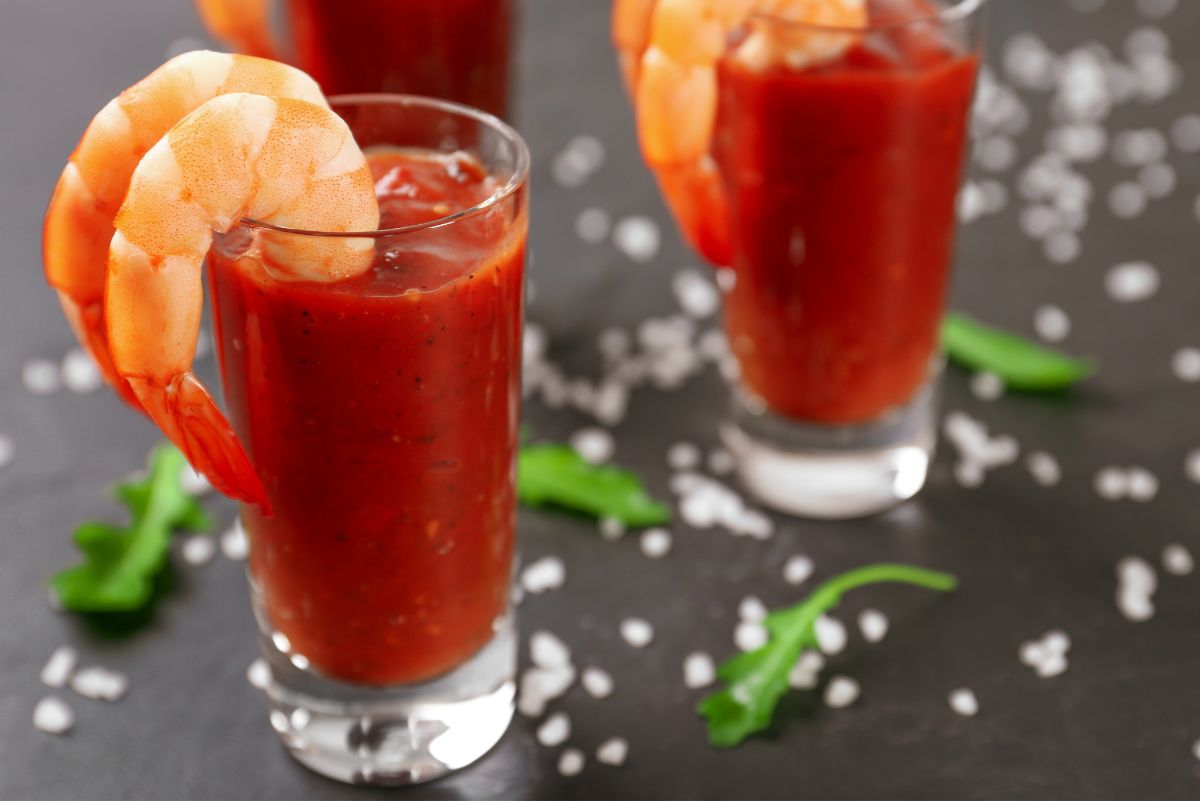 Easy party shrimp cocktail served in tall clear glasses filled with classic red cocktail sauce, each topped with a cooked shrimp hooked over the rim. Set on a dark surface with scattered coarse salt and fresh green herbs, showing a simple shrimp cocktail appetizer ready for serving.