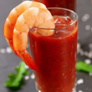 Easy shrimp cocktail appetizer served in a clear glass filled with classic red cocktail sauce, topped with plump cooked shrimp hooked over the rim. Set on a dark surface with scattered salt crystals and fresh green herbs in the background, highlighting a simple WW friendly party appetizer.