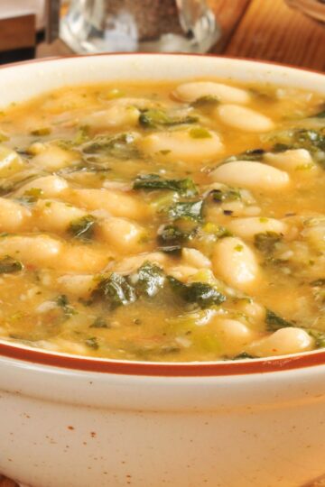 Creamy white bean and kale soup in a ceramic bowl, with tender white beans, chopped kale, and a light golden broth. Hearty vegetable soup with a smooth texture, visible greens, and beans throughout, served warm on a wooden table for a cozy, healthy meal.