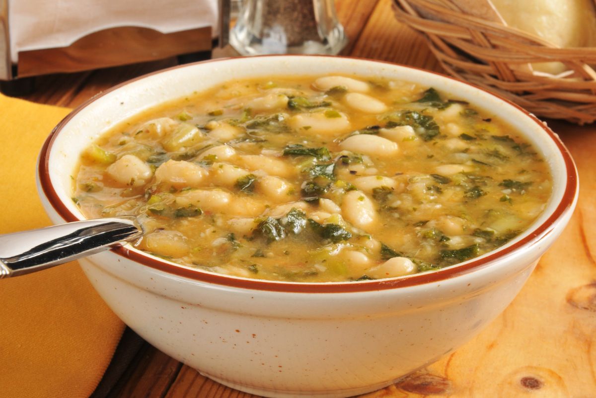 Bowl of white bean and kale soup with a golden vegetable broth, tender white beans, chopped kale, and herbs. A metal spoon rests in the ceramic bowl, set on a wooden table with a napkin, bread basket, and pepper shaker in the background for a cozy, healthy soup meal.