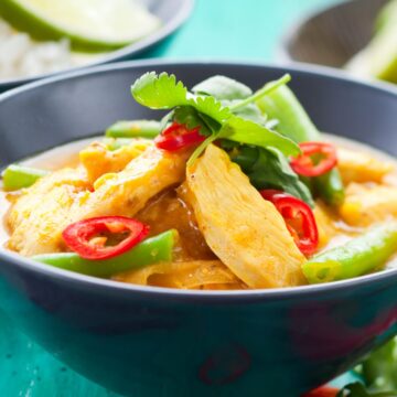 High protein coconut chicken curry in a dark bowl with sliced chicken breast, green bell peppers, and red chili slices in a creamy golden coconut sauce. Topped with fresh cilantro, set on a turquoise wooden table with lime wedges and rice bowls softly blurred in the background.