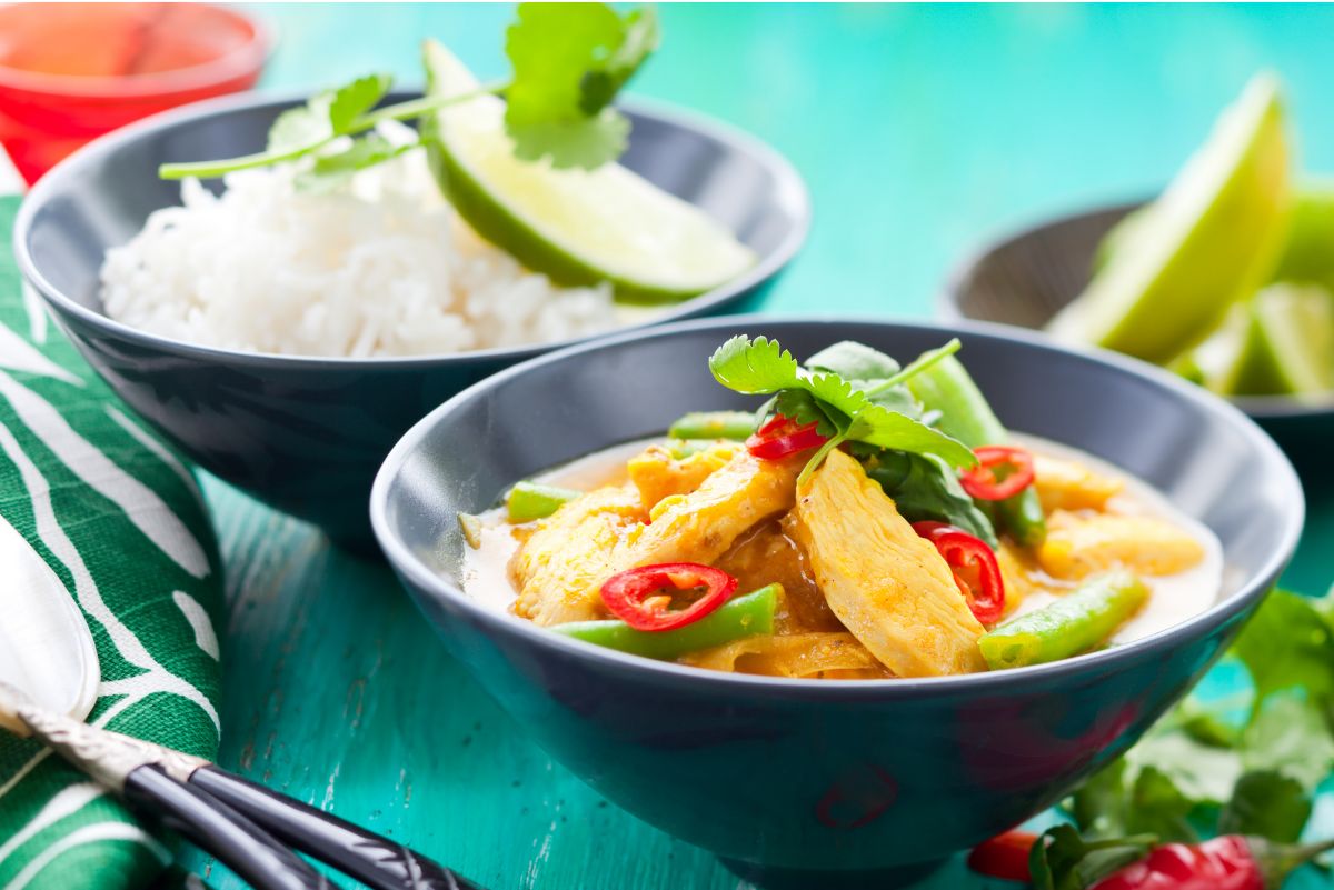 High protein coconut chicken curry served in a dark bowl with sliced chicken breast, green bell peppers, and red chili slices in a creamy coconut sauce. Garnished with fresh cilantro and lime wedges, with a bowl of white rice in the background on a turquoise table.