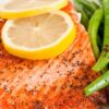 Seasoned baked salmon fillet topped with two lemon slices, served alongside crisp green beans. The salmon is lightly peppered, and both the fish and green beans are presented on a white plate, emphasizing a healthy high protein meal.
