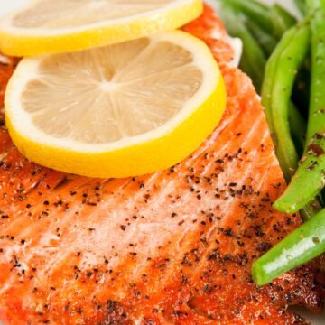 Seasoned baked salmon fillet topped with two lemon slices, served alongside crisp green beans. The salmon is lightly peppered, and both the fish and green beans are presented on a white plate, emphasizing a healthy high protein meal.