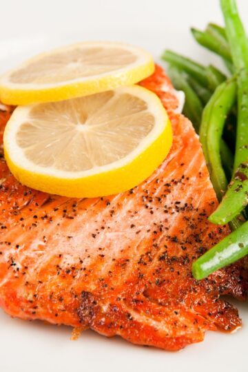 Seasoned baked salmon fillet topped with two lemon slices, served alongside crisp green beans. The salmon is lightly peppered, and both the fish and green beans are presented on a white plate, emphasizing a healthy high protein meal.