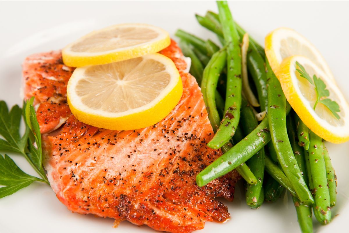 Baked salmon fillet topped with two lemon slices, seasoned with cracked black pepper, served next to green beans and garnished with fresh parsley. A lemon slice with parsley sits on the green beans, all arranged on a white plate for a healthy, high protein meal.
