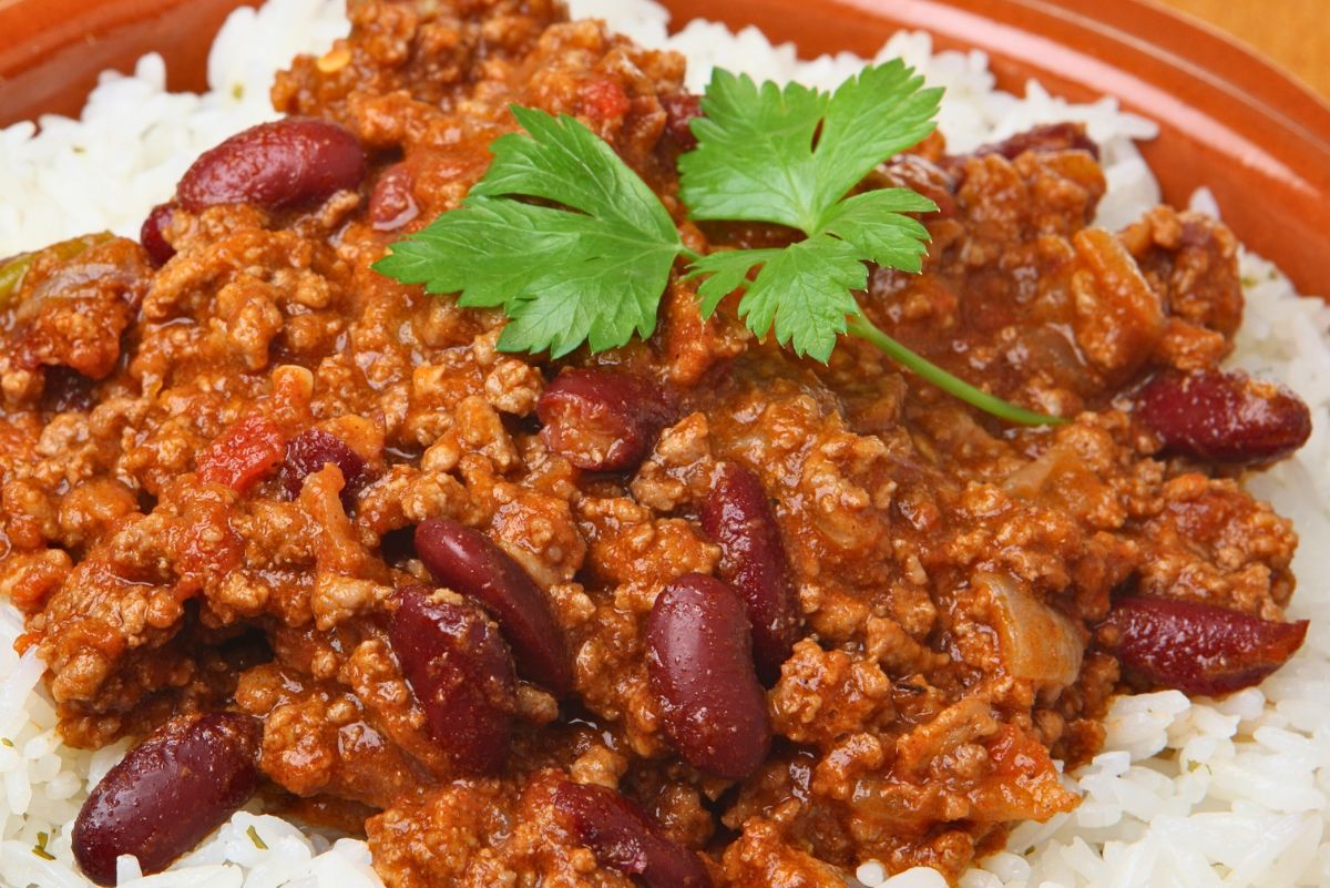 High protein beef chili with ground beef, red kidney beans, and thick tomato sauce served over white rice in a brown bowl. Garnished with fresh green parsley, showing a hearty, saucy texture ideal for an easy weeknight chili dinner.