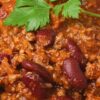 Close-up of high protein beef chili with ground beef, red kidney beans, and thick tomato-based sauce, garnished with fresh green parsley. Hearty, saucy texture with visible beans and beef, served over white rice, highlighting a comforting weeknight chili recipe.